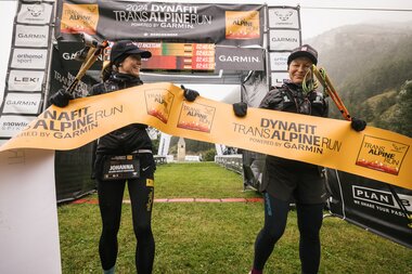 Zwei Läuferinnen jubeln beim Zieleinlauf des Dynafit Transalpine Run 2024, das Zielband in den Händen. | © TAR - Plan B