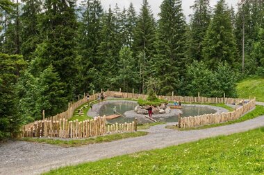 Neuer Spielplatz in Sonntag-Stein | © Seilbahnen Sonntag-Stein Spielplatz am See mit Floß, Balancier-Baumstamm und weiteren Spielstationen, eingebettet in die Natur; vorne eine Wiese, dahinter ein Weg und Bäume im Hintergrund. | © Seilbahnen Sonntag-Stein